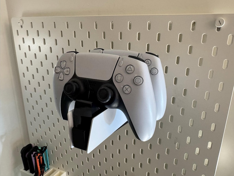 K&ouml;nnte beinhalten: Zwei wei&szlig;e und schwarze Gamecontroller sind an einer wei&szlig;en Lochwand befestigt. Die Controller sind &uuml;bereinander gestapelt, wobei die Kn&ouml;pfe und Joysticks sichtbar sind. Die Lochwand hat ein Raster aus L&ouml;chern.