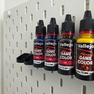以下が含まれることがあります： 青、茶色、黄色など、さまざまな色のVallejo Game Colorインクボトルが白いペグボードに並んでいます。各ボトルには黒いキャップと、ブランド名と色の情報が記載されたラベルが付いています。