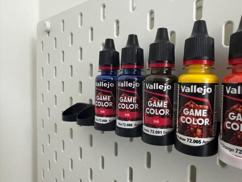 以下が含まれることがあります： 青、茶色、黄色など、さまざまな色のVallejo Game Colorインクボトルが白いペグボードに並んでいます。各ボトルには黒いキャップと、ブランド名と色の情報が記載されたラベルが付いています。