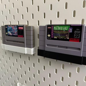 K&ouml;nnte beinhalten: Zwei graue Super Nintendo-Spielmodule, eines mit einem Capcom-Etikett und das andere mit einem PGA Tour Golf-Etikett, an einer wei&szlig;en Lochwand befestigt. Die Module werden von wei&szlig;en und schwarzen Haltern gehalten.