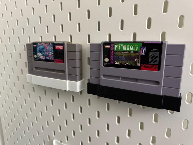 K&ouml;nnte beinhalten: Zwei graue Super Nintendo-Spielmodule, eines mit einem Capcom-Etikett und das andere mit einem PGA Tour Golf-Etikett, an einer wei&szlig;en Lochwand befestigt. Die Module werden von wei&szlig;en und schwarzen Haltern gehalten.