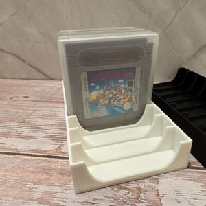 K&ouml;nnte beinhalten: Ein wei&szlig;er 3D-gedruckter Game Boy-Kassettenhalter mit einer Super Mario Land-Spielkassette in einer transparenten Kunststoffh&uuml;lle. Daneben befindet sich ein schwarzer Kassettenhalter. Die Spielkassette hat ein farbenfrohes Etikett.