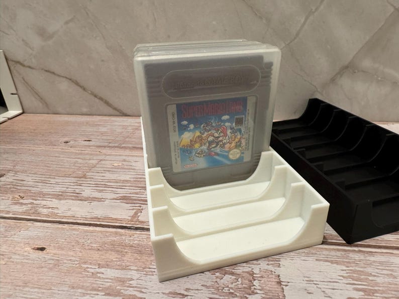 K&ouml;nnte beinhalten: Ein wei&szlig;er 3D-gedruckter Game Boy-Kassettenhalter mit einer Super Mario Land-Spielkassette in einer transparenten Kunststoffh&uuml;lle. Daneben befindet sich ein schwarzer Kassettenhalter. Die Spielkassette hat ein farbenfrohes Etikett.