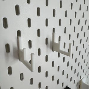 Puede incluir: Panel perforado blanco con orificios ovalados espaciados. Dos ganchos de pl&aacute;stico blanco est&aacute;n insertados en el panel. Los ganchos est&aacute;n dise&ntilde;ados para colgar objetos. El panel perforado es una soluci&oacute;n de almacenamiento vers&aacute;til.