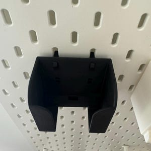 May include: A black plastic pegboard bin with a curved front and two small tabs on the top. The bin is mounted on a white pegboard with evenly spaced holes. The bin is designed to hold small items.