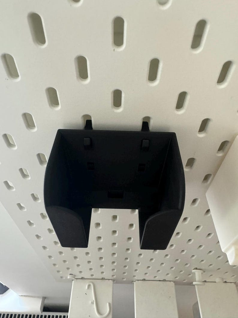 May include: A black plastic pegboard bin with a curved front and two small tabs on the top. The bin is mounted on a white pegboard with evenly spaced holes. The bin is designed to hold small items.