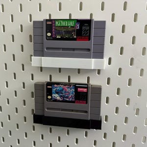 K&ouml;nnte beinhalten: Zwei graue Super Nintendo-Spielmodule, eines mit der Aufschrift "PGA TOUR GOLF" und das andere mit "STREET FIGHTER II". Die Module sind an einer wei&szlig;en Lochwand befestigt.
