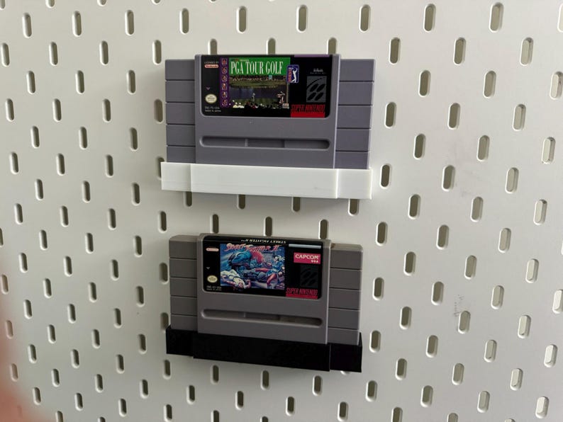 K&ouml;nnte beinhalten: Zwei graue Super Nintendo-Spielmodule, eines mit der Aufschrift "PGA TOUR GOLF" und das andere mit "STREET FIGHTER II". Die Module sind an einer wei&szlig;en Lochwand befestigt.