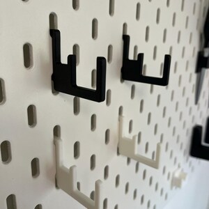 Puede incluir: Primer plano de un tablero perforado blanco con varios ganchos negros y blancos insertados. Los ganchos est&aacute;n dise&ntilde;ados para sujetar objetos y est&aacute;n hechos de un material resistente. El tablero tiene agujeros espaciados.