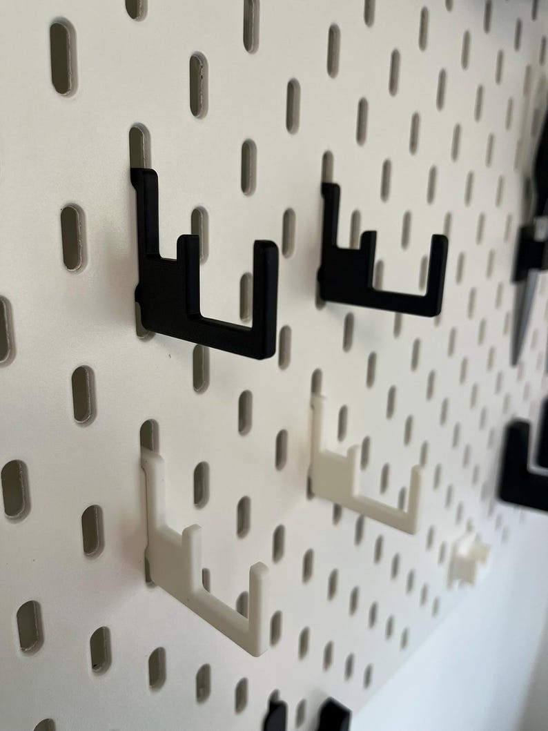 Puede incluir: Primer plano de un tablero perforado blanco con varios ganchos negros y blancos insertados. Los ganchos est&aacute;n dise&ntilde;ados para sujetar objetos y est&aacute;n hechos de un material resistente. El tablero tiene agujeros espaciados.