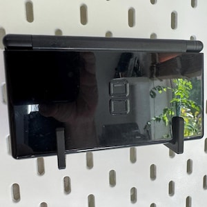 Puede incluir: Consola de juegos port&aacute;til negra brillante, montada en una pared perforada blanca. La consola se sujeta con dos soportes negros. La pantalla refleja una ventana y vegetaci&oacute;n cercana.