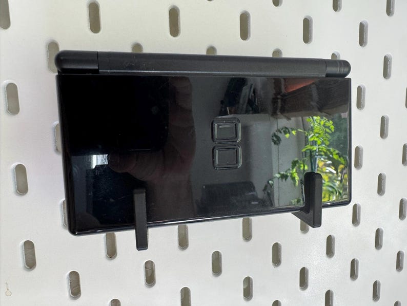 Puede incluir: Consola de juegos port&aacute;til negra brillante, montada en una pared perforada blanca. La consola se sujeta con dos soportes negros. La pantalla refleja una ventana y vegetaci&oacute;n cercana.