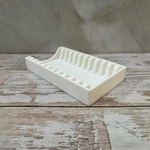 Op de afbeelding: Een witte rechthoekige zeepbakje met een geribbeld ontwerp. Het bakje heeft een verhoogde achterkant en een reeks verticale ribbels aan &eacute;&eacute;n kant. Het zeepbakje is gemaakt van een solide, ondoorzichtig materiaal.