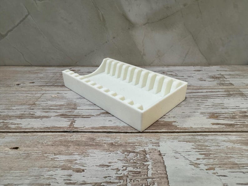 Op de afbeelding: Een witte rechthoekige zeepbakje met een geribbeld ontwerp. Het bakje heeft een verhoogde achterkant en een reeks verticale ribbels aan &eacute;&eacute;n kant. Het zeepbakje is gemaakt van een solide, ondoorzichtig materiaal.