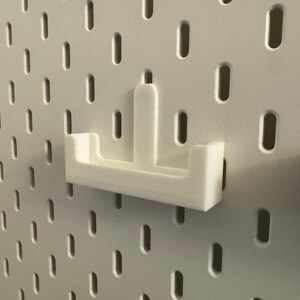 以下が含まれることがあります： 白の3Dプリントされたペグボードシェルフアタッチメント。シェルフは、隆起した背面と2つのサイドサポートを備えた長方形のベースを備えています。楕円形の穴のある白いペグボードに取り付けるように設計されています。黒いペグボードアタッチメントが見えます。