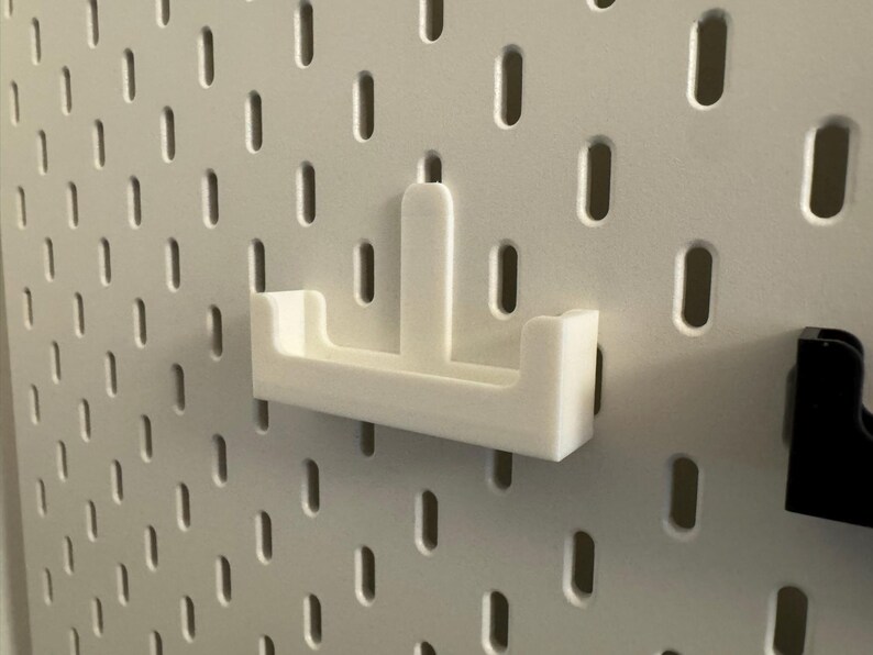 以下が含まれることがあります： 白の3Dプリントされたペグボードシェルフアタッチメント。シェルフは、隆起した背面と2つのサイドサポートを備えた長方形のベースを備えています。楕円形の穴のある白いペグボードに取り付けるように設計されています。黒いペグボードアタッチメントが見えます。