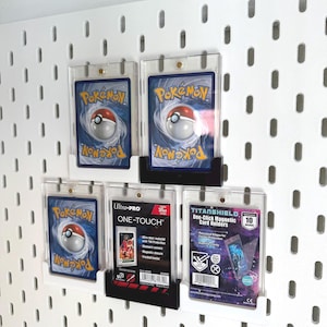 May include: A collection of Pokémon trading cards displayed in clear protective cases, mounted on a white pegboard. The cards feature the Pokémon logo and a Poké Ball design. Includes Ultra-PRO and TITANSHIELD card holders.