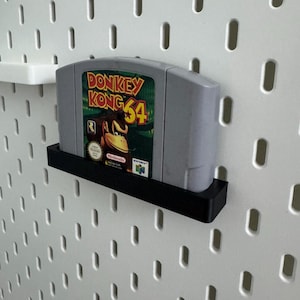 May include: A gray Donkey Kong 64 video game cartridge is displayed on a black shelf against a white pegboard. The game's label features Donkey Kong and the text "Donkey Kong 64". The shelf is mounted on a white pegboard.