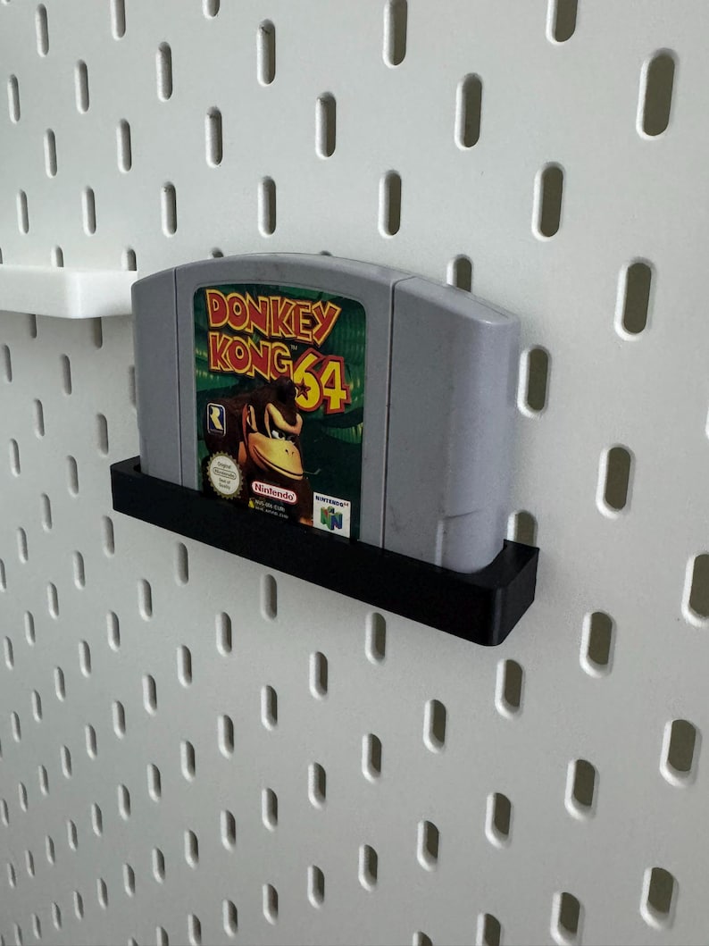 May include: A gray Donkey Kong 64 video game cartridge is displayed on a black shelf against a white pegboard. The game's label features Donkey Kong and the text "Donkey Kong 64". The shelf is mounted on a white pegboard.
