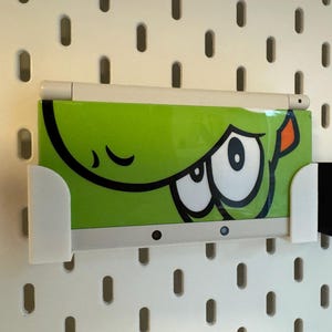 Puede incluir: Un dispositivo electr&oacute;nico verde con un dise&ntilde;o de ojo de dibujos animados se sostiene en un soporte blanco. Un l&aacute;piz &oacute;ptico blanco descansa encima. El soporte est&aacute; montado en una placa perforada blanca.