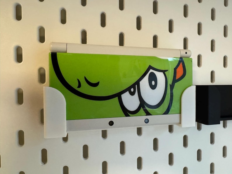 Puede incluir: Un dispositivo electr&oacute;nico verde con un dise&ntilde;o de ojo de dibujos animados se sostiene en un soporte blanco. Un l&aacute;piz &oacute;ptico blanco descansa encima. El soporte est&aacute; montado en una placa perforada blanca.