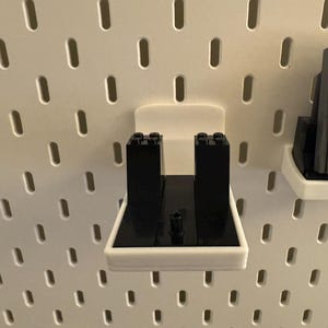May include: White 3D-printed pegboard accessories. One holder features black Lego bricks, and another holds a Nintendo Game Boy cartridge. The pegboard has evenly spaced oval holes.