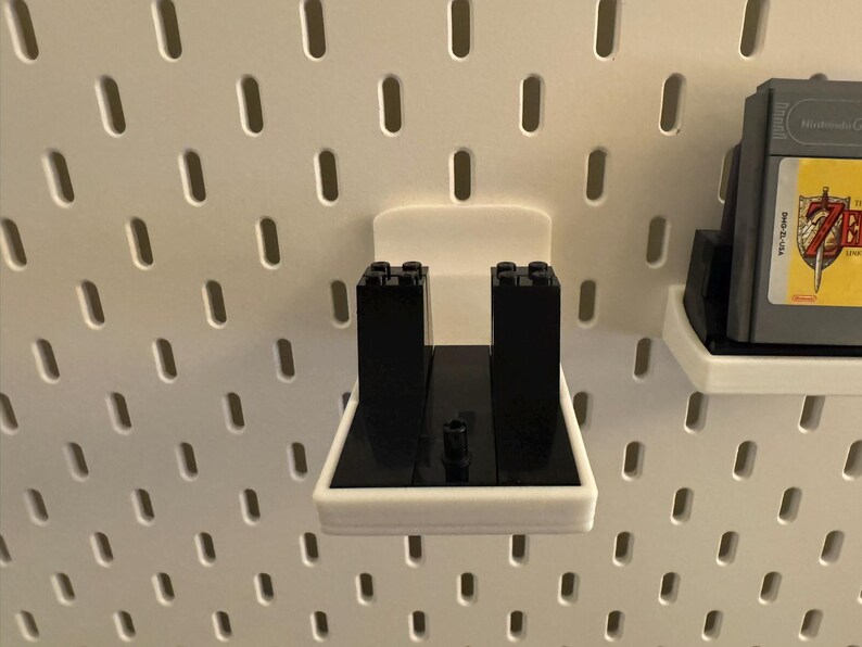 May include: White 3D-printed pegboard accessories. One holder features black Lego bricks, and another holds a Nintendo Game Boy cartridge. The pegboard has evenly spaced oval holes.