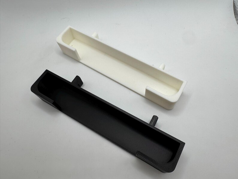 May include: Two rectangular, open-top containers. One is black, the other is white. Both have a similar design with a long, narrow shape and a lip on each end. The black container has a small, cylindrical protrusion on one side.