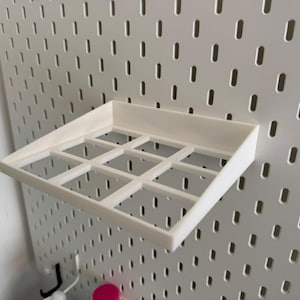 Op de afbeelding: Wit plasticen plank met negen vierkante vakken, gemonteerd op een witte prikbordwand.