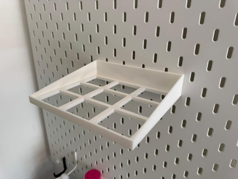 Op de afbeelding: Wit plasticen plank met negen vierkante vakken, gemonteerd op een witte prikbordwand.