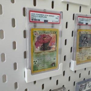 Peut inclure: Carte Pok&eacute;mon grad&eacute;e Erika's Vileplume de l'ensemble Gym Heroes 2000. La carte est dans un &eacute;tui de protection et expos&eacute;e sur un panneau perfor&eacute; blanc. L'illustration de la carte repr&eacute;sente une cr&eacute;ature v&eacute;g&eacute;tale.