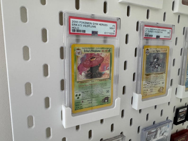 Peut inclure: Carte Pok&eacute;mon grad&eacute;e Erika's Vileplume de l'ensemble Gym Heroes 2000. La carte est dans un &eacute;tui de protection et expos&eacute;e sur un panneau perfor&eacute; blanc. L'illustration de la carte repr&eacute;sente une cr&eacute;ature v&eacute;g&eacute;tale.