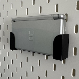 Op de afbeelding: Een zilveren Nintendo DS handheld-spelconsole is gemonteerd op een zwarte plastic wandbevestiging. De bevestiging is bevestigd aan een witte pegboard-wand.