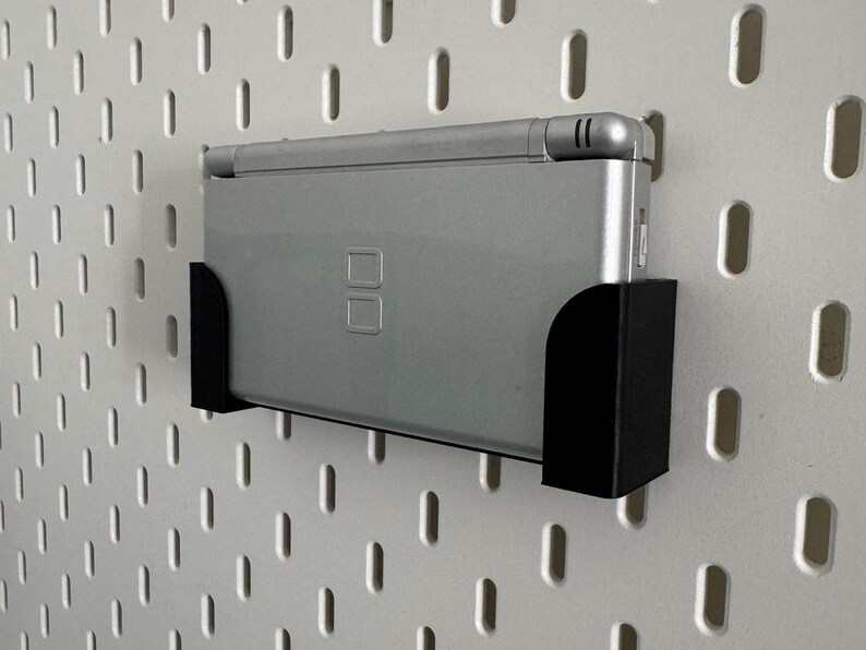 Op de afbeelding: Een zilveren Nintendo DS handheld-spelconsole is gemonteerd op een zwarte plastic wandbevestiging. De bevestiging is bevestigd aan een witte pegboard-wand.
