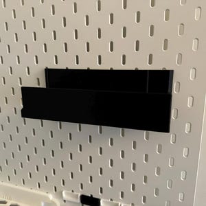 Peut inclure: Une &eacute;tag&egrave;re de rangement noire et rectangulaire fix&eacute;e sur un panneau perfor&eacute; blanc. L'&eacute;tag&egrave;re est faite d'un mat&eacute;riau opaque et solide et est con&ccedil;ue pour contenir de petits objets. Le panneau perfor&eacute; a une grille de trous espac&eacute;s.