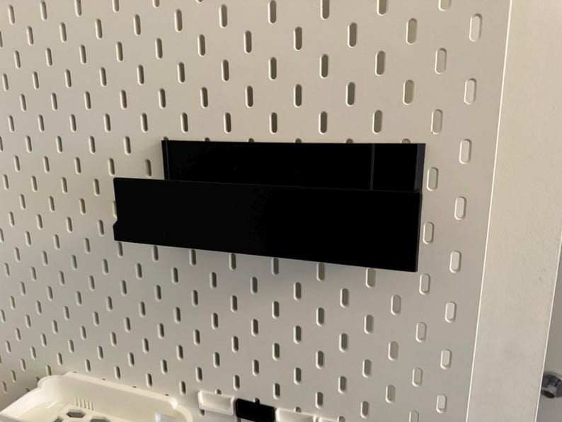 Peut inclure: Une &eacute;tag&egrave;re de rangement noire et rectangulaire fix&eacute;e sur un panneau perfor&eacute; blanc. L'&eacute;tag&egrave;re est faite d'un mat&eacute;riau opaque et solide et est con&ccedil;ue pour contenir de petits objets. Le panneau perfor&eacute; a une grille de trous espac&eacute;s.