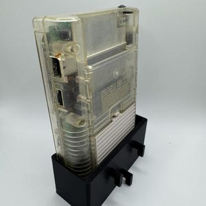 Op de afbeelding: Een transparante, doorzichtige Game Boy Color handheld-videogamesysteem wordt getoond in een zwarte plastic standaard. De console heeft een zwarte aan/uit-knop en een zwarte cartridge-sleuf. De standaard heeft twee kleine zwarte lipjes aan de zijkanten.