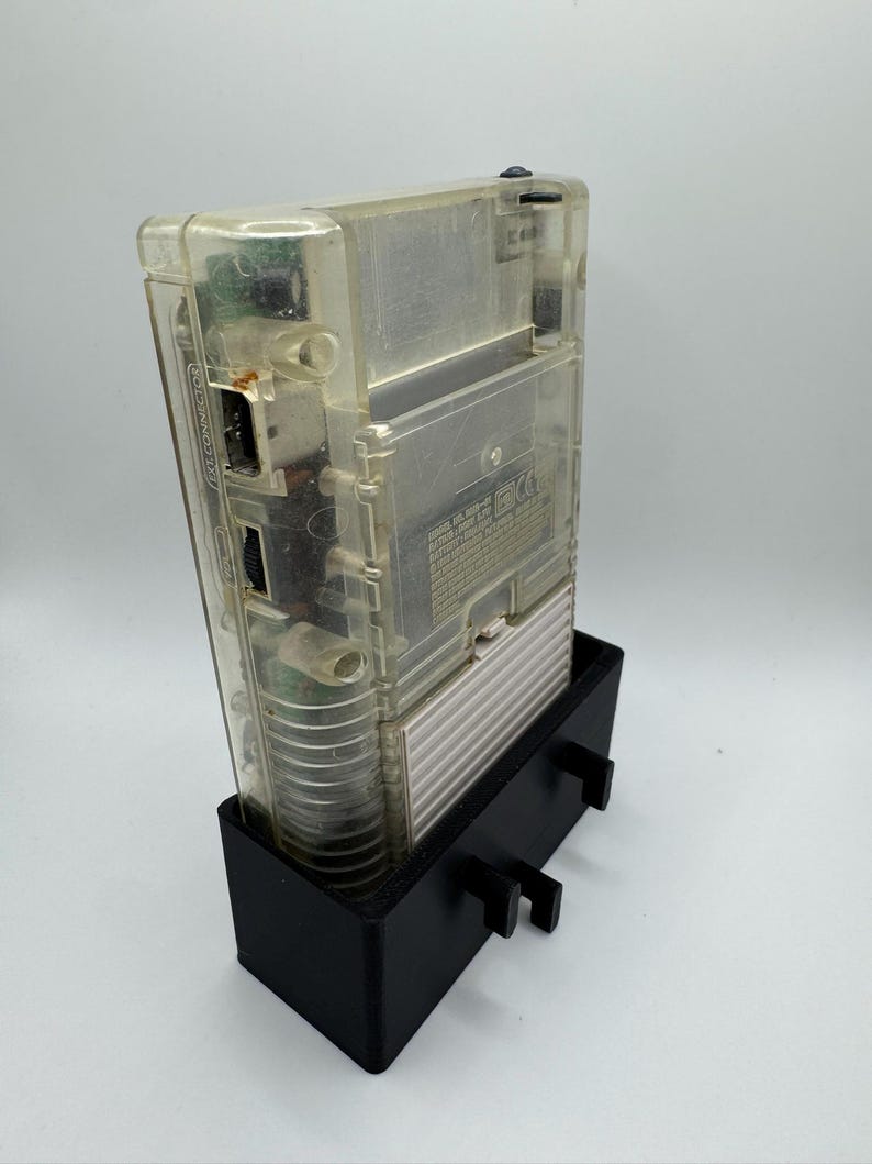 Op de afbeelding: Een transparante, doorzichtige Game Boy Color handheld-videogamesysteem wordt getoond in een zwarte plastic standaard. De console heeft een zwarte aan/uit-knop en een zwarte cartridge-sleuf. De standaard heeft twee kleine zwarte lipjes aan de zijkanten.