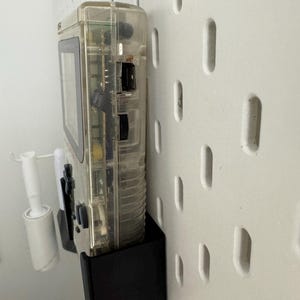 Op de afbeelding: Een transparante, doorschijnende handheld-videogames console met een zwart-wit scherm en een zwarte plastic standaard. De console is gemonteerd op een witte pegboard-wand.