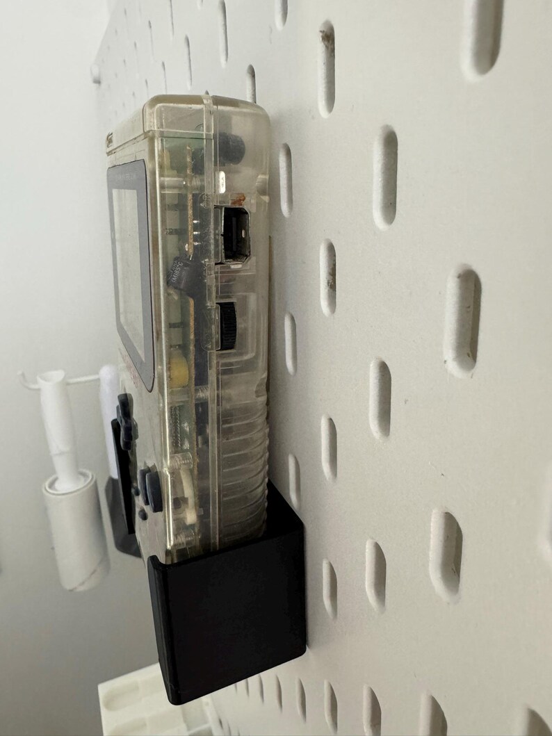 Op de afbeelding: Een transparante, doorschijnende handheld-videogames console met een zwart-wit scherm en een zwarte plastic standaard. De console is gemonteerd op een witte pegboard-wand.