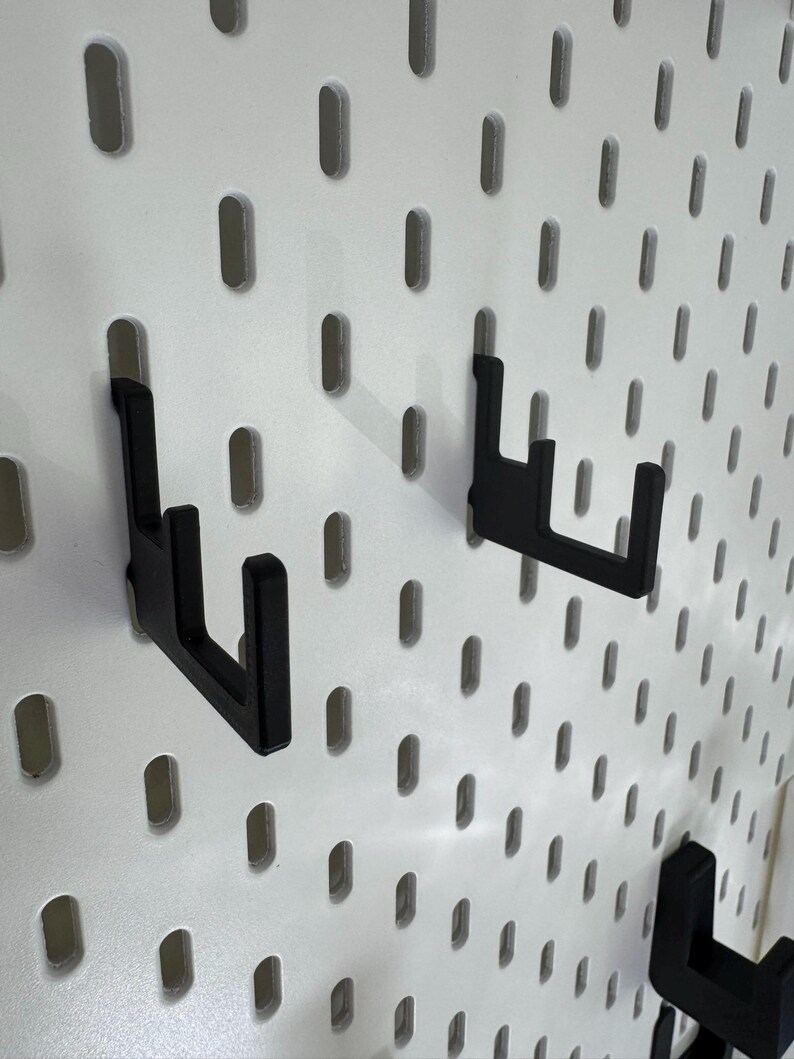 Puede incluir: Ganchos negros para panel perforado fijados a un panel perforado blanco. Los ganchos son de material oscuro y est&aacute;n dise&ntilde;ados para sujetar objetos. El panel perforado tiene agujeros ovalados espaciados.