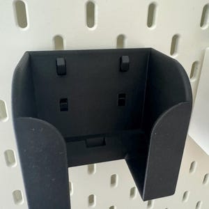 May include: A black, wall-mounted holder with a U-shaped design. The holder has a rectangular base and two side supports. It features two small, rectangular protrusions on the top and two on the front. The holder is designed to be mounted on a pegboard.