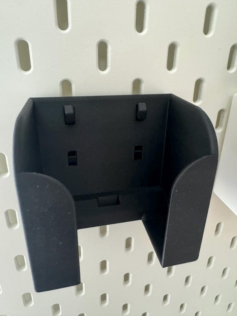 May include: A black, wall-mounted holder with a U-shaped design. The holder has a rectangular base and two side supports. It features two small, rectangular protrusions on the top and two on the front. The holder is designed to be mounted on a pegboard.