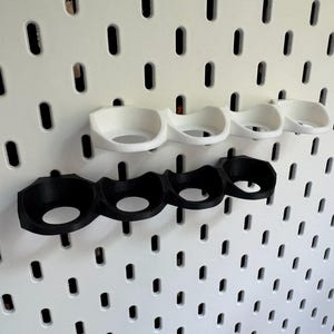 Puede incluir: Dos juegos de portavasos, uno negro y uno blanco, montados en una pared de tablero de clavijas. Cada juego tiene cuatro portavasos.