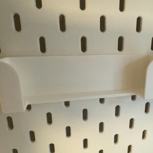 Puede incluir: Un estante blanco rectangular con bordes curvos, dise&ntilde;ado para fijarse a una panel perforado. El estante est&aacute; hecho de un material s&oacute;lido y opaco y est&aacute; montado en un panel perforado blanco con agujeros ovalados espaciados uniformemente. Un contenedor negro est&aacute; adjunto a la derecha.