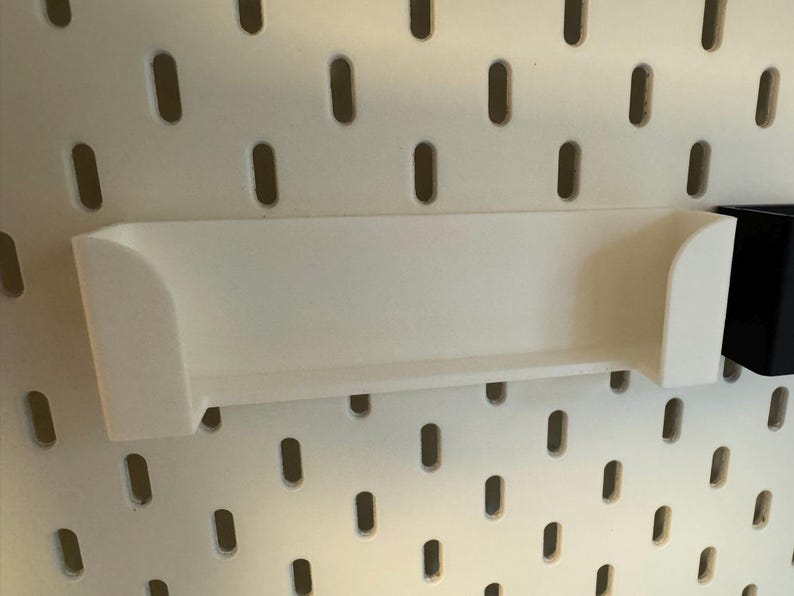 Puede incluir: Un estante blanco rectangular con bordes curvos, dise&ntilde;ado para fijarse a una panel perforado. El estante est&aacute; hecho de un material s&oacute;lido y opaco y est&aacute; montado en un panel perforado blanco con agujeros ovalados espaciados uniformemente. Un contenedor negro est&aacute; adjunto a la derecha.