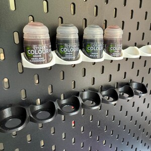以下が含まれることがあります： 黒色の蓋と「Citadel Colour」と色合いの名前が書かれたラベルが付いた小さな塗料瓶が5本セットになっています。瓶は黒いフックが付いた白いラックに掛けられています。ラックは黒いペグボードの壁に取り付けられています。