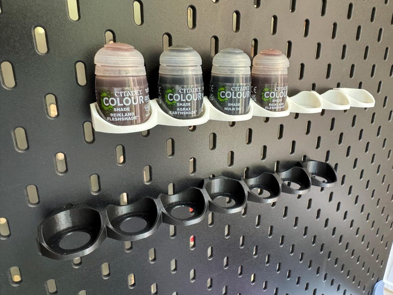 以下が含まれることがあります： 黒色の蓋と「Citadel Colour」と色合いの名前が書かれたラベルが付いた小さな塗料瓶が5本セットになっています。瓶は黒いフックが付いた白いラックに掛けられています。ラックは黒いペグボードの壁に取り付けられています。