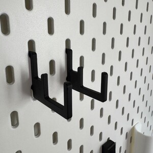 May include: Black metal hooks on a white pegboard. The hooks fit into the pegboard's oval holes, designed for storage. The pegboard is a storage and organisation system, ideal for various items.