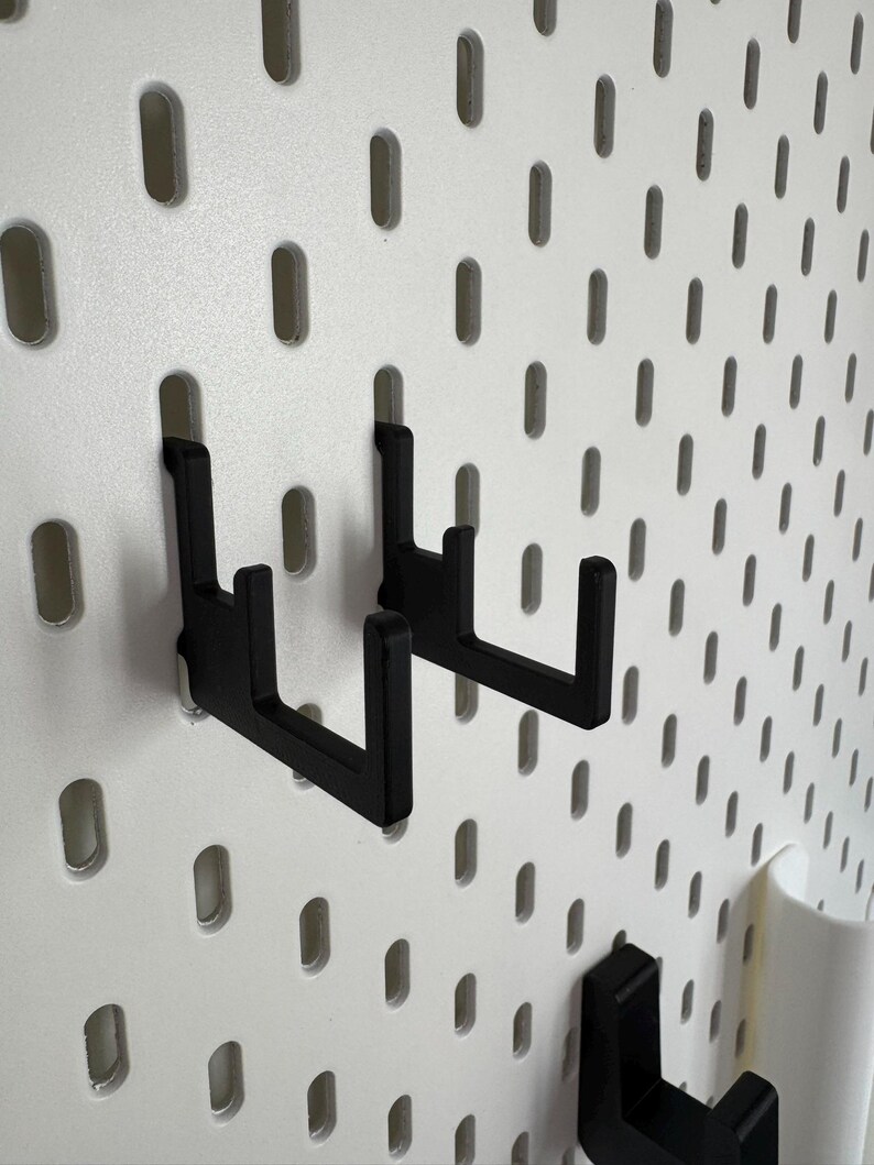 May include: Black metal hooks on a white pegboard. The hooks fit into the pegboard's oval holes, designed for storage. The pegboard is a storage and organisation system, ideal for various items.